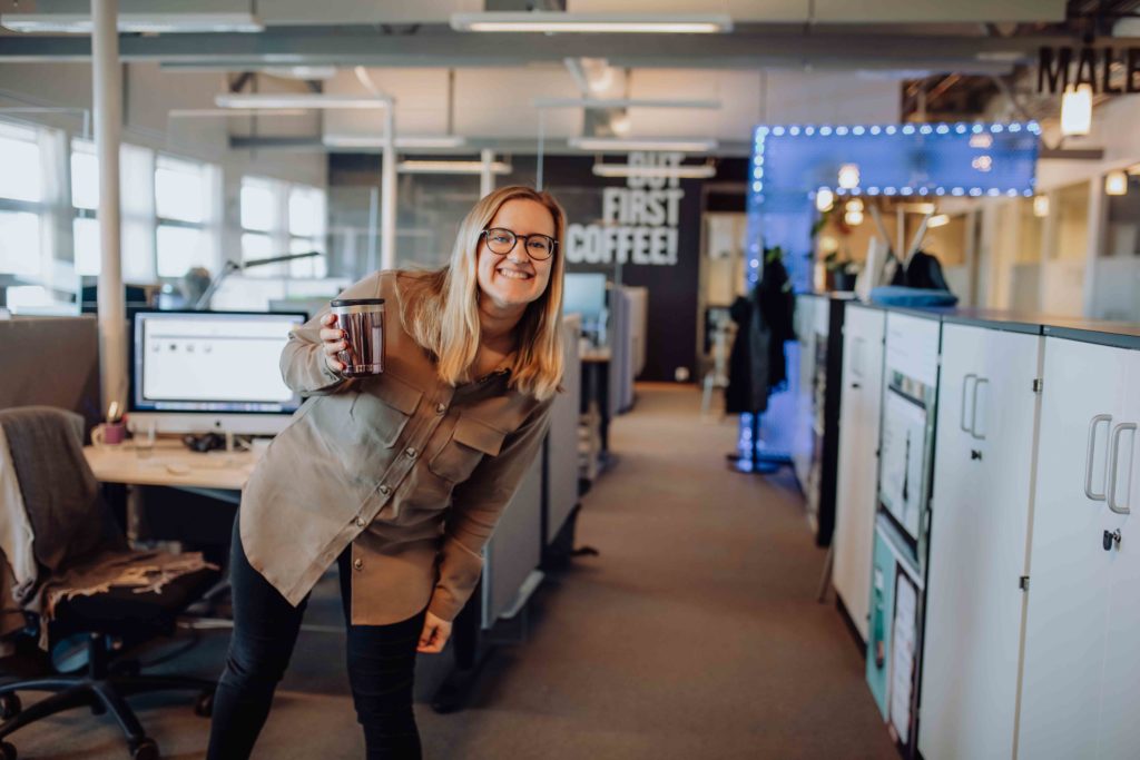 En kvinne med briller holder en kaffekopp og lener seg smilende mot fotografen. I bakgrunnen ses et åpent kontorlandskap.