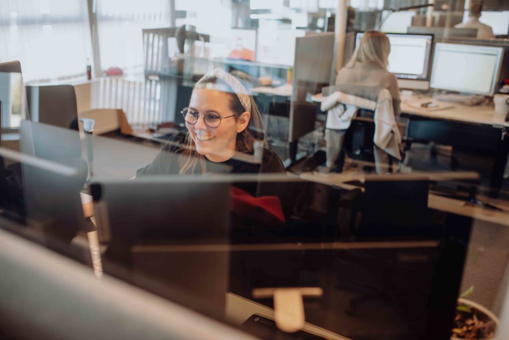 En kvinne med briller sitter ved arbeidsplassen sin og smiler. Bildet er tatt gjennom et plexiglass og i bakgrunnen ses andre som også sitter ved sine arbeidsplasser.