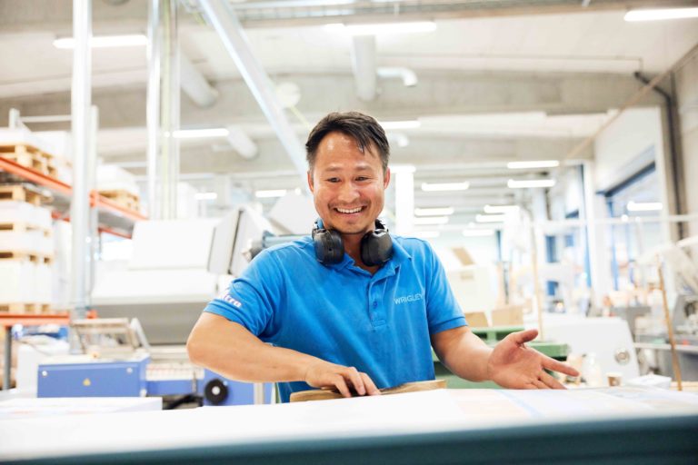 En smilende mann i et produksjonslokale. Mannen har på blå t-skjorte og har hodetelefoner rundt halsen mens han arbeider.